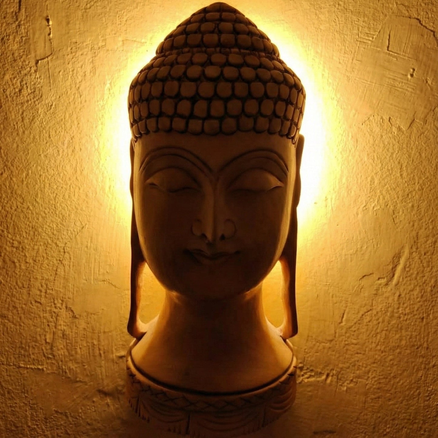 A detailed front view of a hand-carved light wood Buddha head statue with a serene expression, closed eyes for meditation, and intricately carved hair details against a solid dark blue background.
