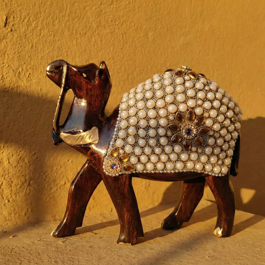 A hand-carved dark wooden camel figurine standing in profile, decorated with an ornate saddle made of white pearls and colored stones, set against a warm yellow background