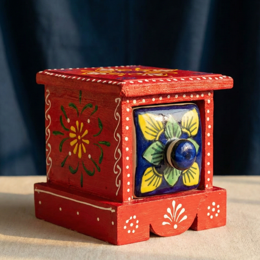A close-up photograph of a handcrafted red wooden chest decorated with white traditional patterns, featuring a square, hand-painted ceramic drawer with green, blue, and yellow floral designs.
