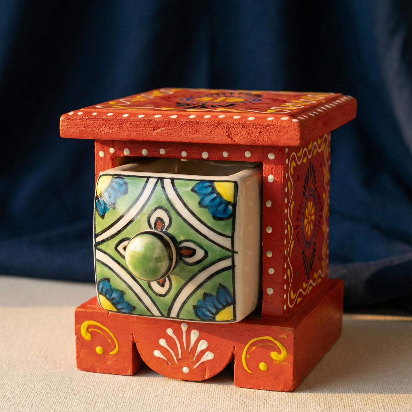 Studio shot of an orange  handcrafted wooden chest featuring a square ceramic drawer with intricate green and blue floral patterns, isolated on a dark blue background.