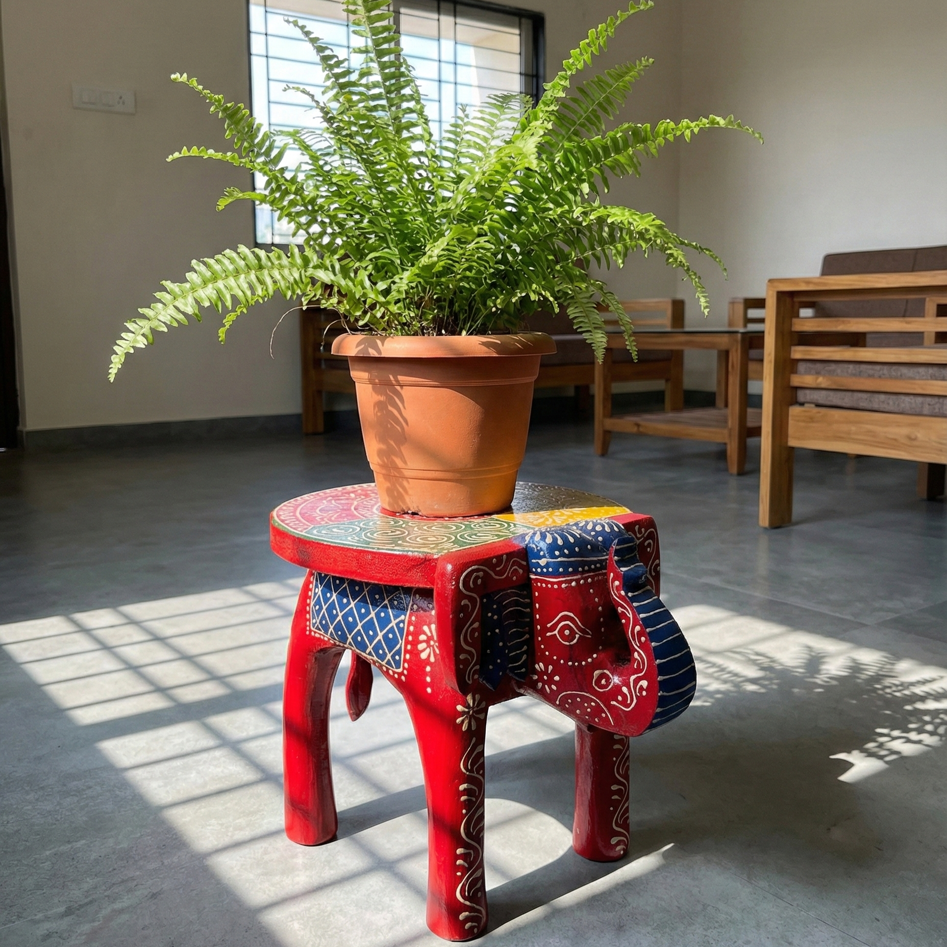 Red hand-painted wooden elephant stool used as a decorative plant stand in a sunlit living room. Ethnic Rajasthani low stool holding a potted fern
