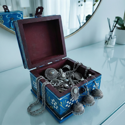 Open blue hand-painted wooden jewelry box filled with silver rings, necklaces, and traditional Indian jhumka earrings, serving as a decorative organizer on a dresser.