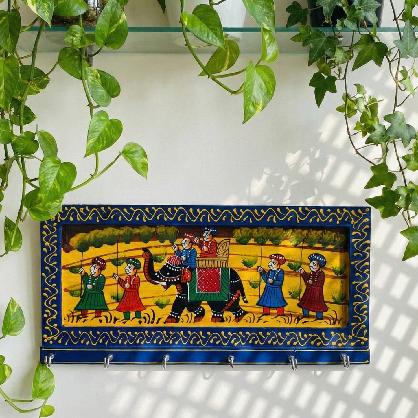 A handcrafted blue wooden key holder for the wall featuring traditional Rajasthani elephant painting, hanging in an entryway surrounded by green plants.