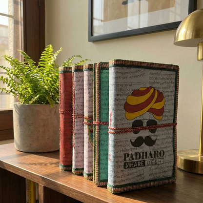 Row of handmade diaries standing on a shelf, highlighting the 'Padharo Mhare Desh' Rajasthani design and unique spine stitching.