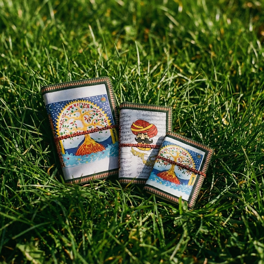 Set of 3 handmade eco-friendly notebooks featuring Tree of Life and Rajasthani Man covers. Styled on green grass to showcase natural recycled cotton paper.
