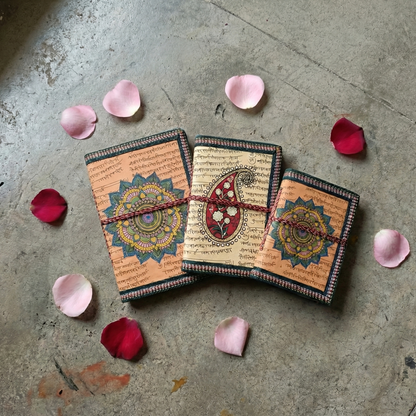 Set of 3 beige handmade eco-friendly notebooks featuring Mandala and Paisley art covers. Flat lay styling with pink rose petals on a rustic stone surface
