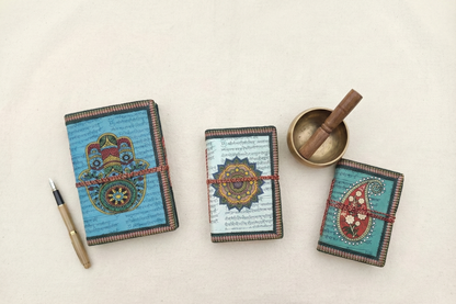 Set of 3 handmade paper notebooks featuring Hamsa, Mandala, and Paisley art covers. Flat lay on white background with fountain pen and singing bowl for spiritual stationery