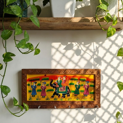 A handcrafted golden brown wooden key holder for the wall featuring traditional Rajasthani elephant painting, hanging in an entryway surrounded by green plants.