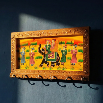 handpainted-wooden-key-hanger-golden-ethnic-design with a painting of people on an elephant, framed in gold, mounted on a dark blue wall.