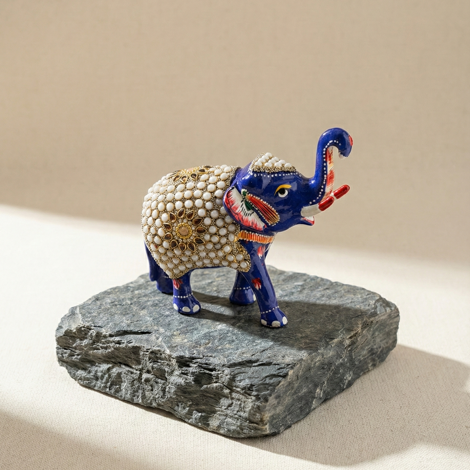 Side profile of a handcrafted blue elephant idol perched on a natural grey stone slab, showcasing the vibrant enamel paint and gold detailing.