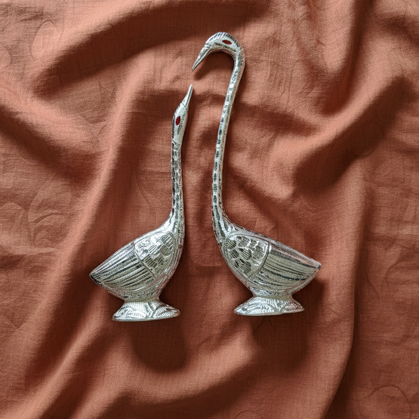 Pair of elegant white metal swan figurines with an antique silver finish, placed on a rust-colored fabric background.