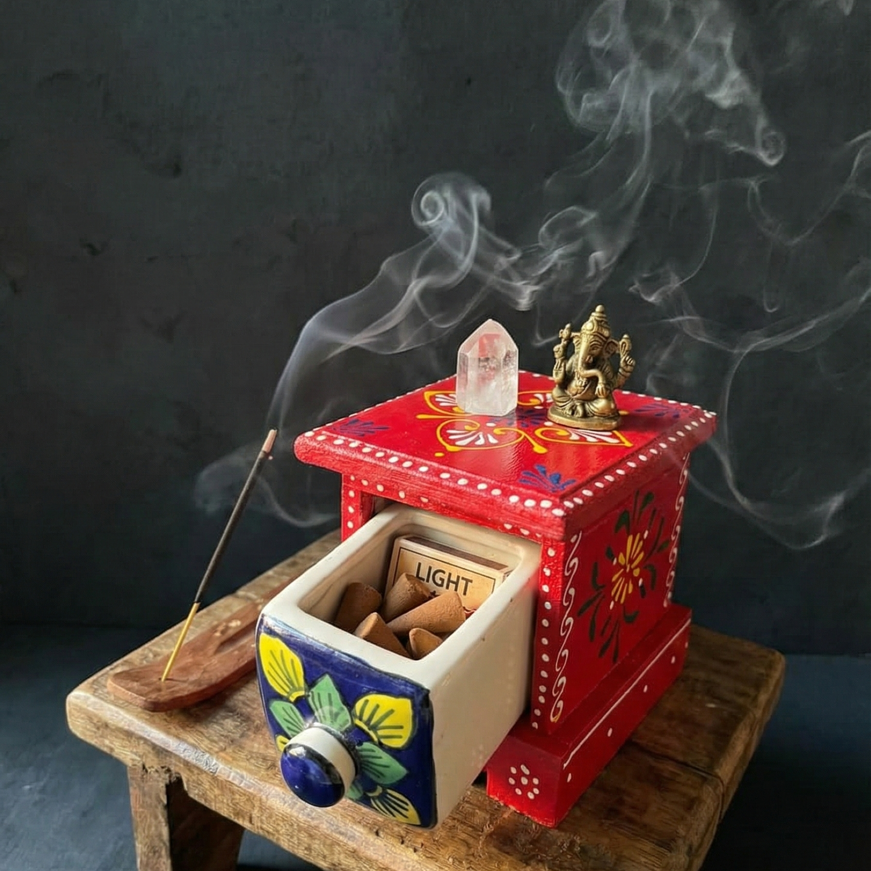 Red wooden chest with ceramic drawer used to store incense cones, placed on a meditation altar with a Ganesha statue and crystal, surrounded by swirling incense smoke.