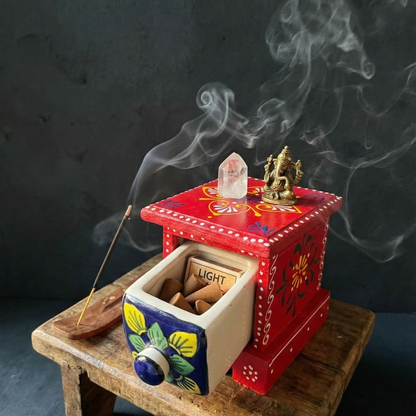 Red wooden chest with ceramic drawer used to store incense cones, placed on a meditation altar with a Ganesha statue and crystal, surrounded by swirling incense smoke.