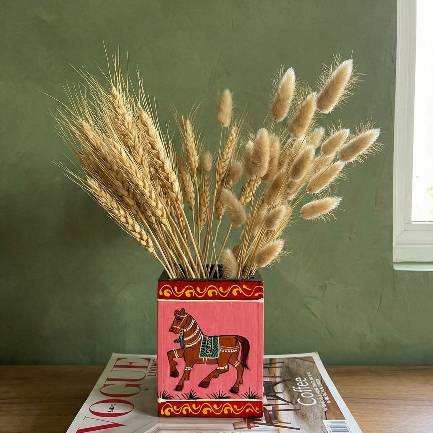 Wooden Pink colour handpainted horse design vase with dried plants on top of magazines against a green wall.