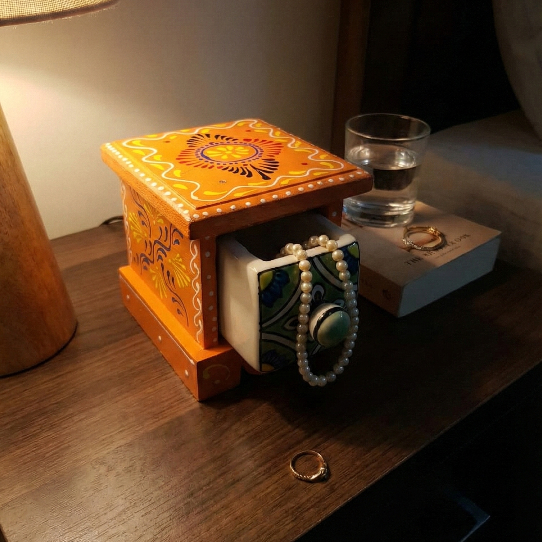 Small wooden chest placed on a bedside nightstand under a lamp, with the ceramic drawer open holding a pearl necklace, serving as an elegant jewelry organizer and ring holder.